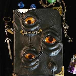 May include: A black leather-bound book with three orange and yellow eyes embossed on the cover. The book has brass corner protectors and a small chain. Additional items include a sword keychain, dice, and a green pendant.