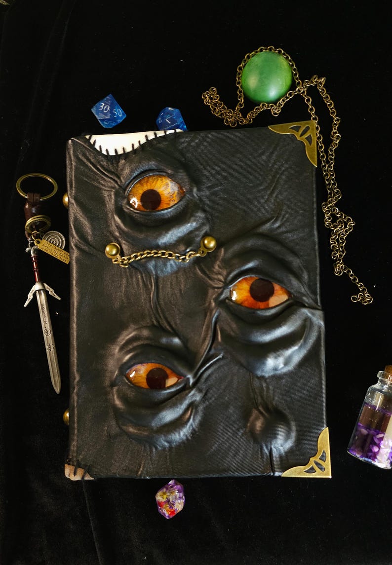 May include: A black leather-bound book with three orange and yellow eyes embossed on the cover. The book has brass corner protectors and a small chain. Additional items include a sword keychain, dice, and a green pendant.