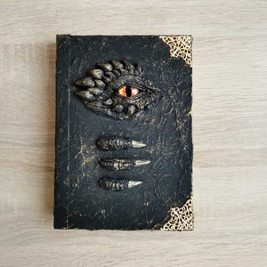 Op de afbeelding: Een zwart lederen gebonden dagboek met een drakenoog en klauwen die op de voorzijde zijn gegraveerd. De omslag heeft gouden accenten en een textuur.