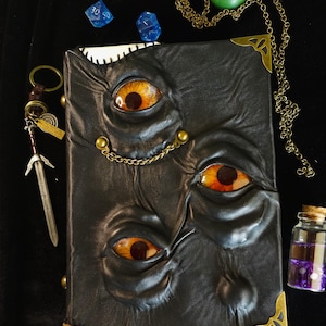 May include: A black leather-bound book with three sets of orange and yellow eyes embossed on the cover. The book is adorned with brass corner protectors and a chain. Additional items include dice, a pendant, a miniature sword, and a small vial.