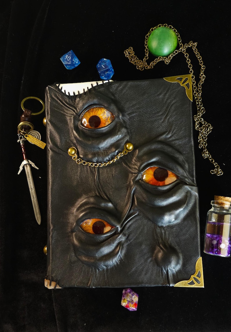 May include: A black leather-bound book with three sets of orange and yellow eyes embossed on the cover. The book is adorned with brass corner protectors and a chain. Additional items include dice, a pendant, a miniature sword, and a small vial.