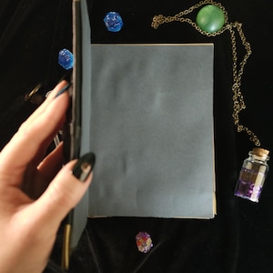 May include: A blue-grey journal is held open, revealing blank pages. A gold-colored chain with a green stone pendant and a small glass bottle with purple contents are next to the journal. Scattered around are blue dice and a crystal.
