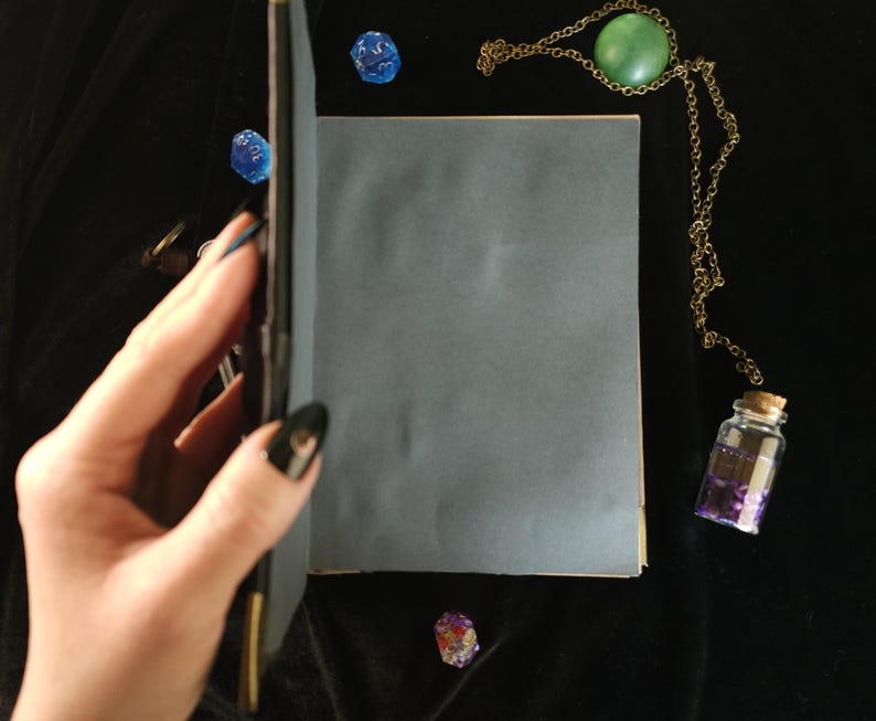 May include: A blue-grey journal is held open, revealing blank pages. A gold-colored chain with a green stone pendant and a small glass bottle with purple contents are next to the journal. Scattered around are blue dice and a crystal.
