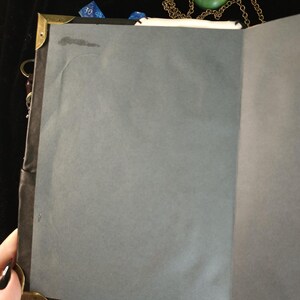 May include: An open journal with gray pages, brass corner protectors, and a black spine. A green stone pendant on a gold chain rests on top. Blue dice are visible near the top edge, suggesting a theme of gaming or fantasy.