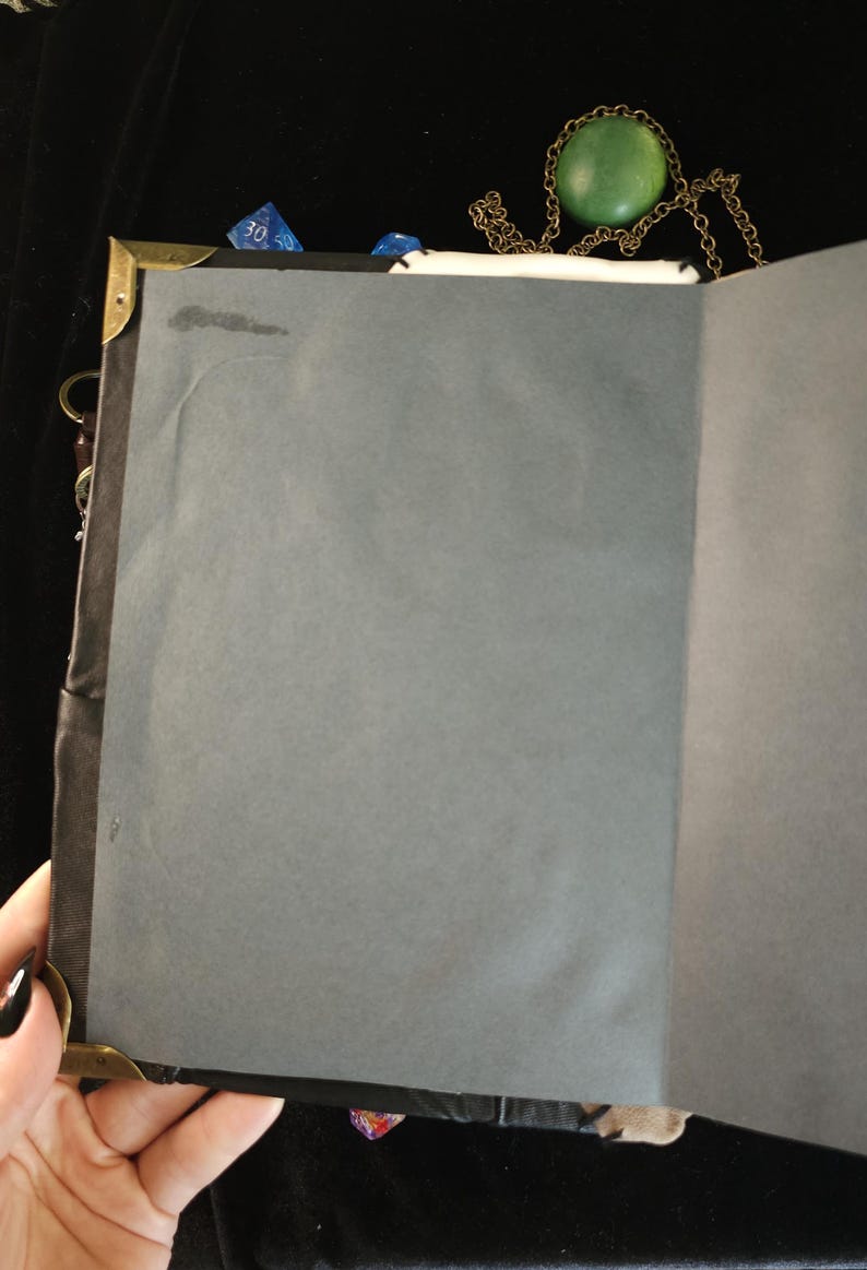 May include: An open journal with gray pages, brass corner protectors, and a black spine. A green stone pendant on a gold chain rests on top. Blue dice are visible near the top edge, suggesting a theme of gaming or fantasy.