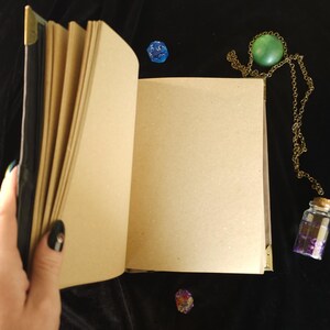 May include: An open, blank journal with kraft paper pages, held by a hand with a black nail. A green stone pendant on a gold chain, a small glass bottle with a cork, and two dice are on a black surface.