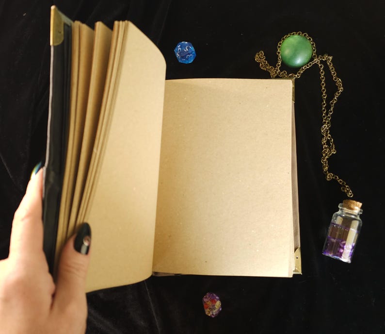 May include: An open, blank journal with kraft paper pages, held by a hand with a black nail. A green stone pendant on a gold chain, a small glass bottle with a cork, and two dice are on a black surface.