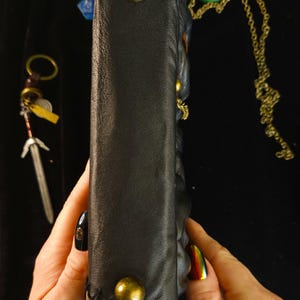 May include: Black leather-bound journal with brass studs and a stitched leather edge. A small sword charm, a green pendant necklace, and a blue die are visible. The journal is held by a person.