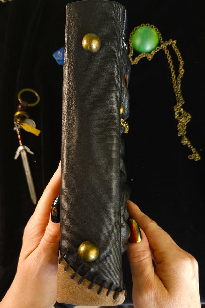 May include: Black leather-bound journal with brass studs and a stitched leather edge. A small sword charm, a green pendant necklace, and a blue die are visible. The journal is held by a person.