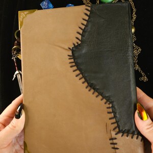 May include: A handmade leather journal with a two-tone design. The journal features a tan cover with a black leather accent stitched together with black thread. Decorative metal corner pieces and a sword charm are visible.