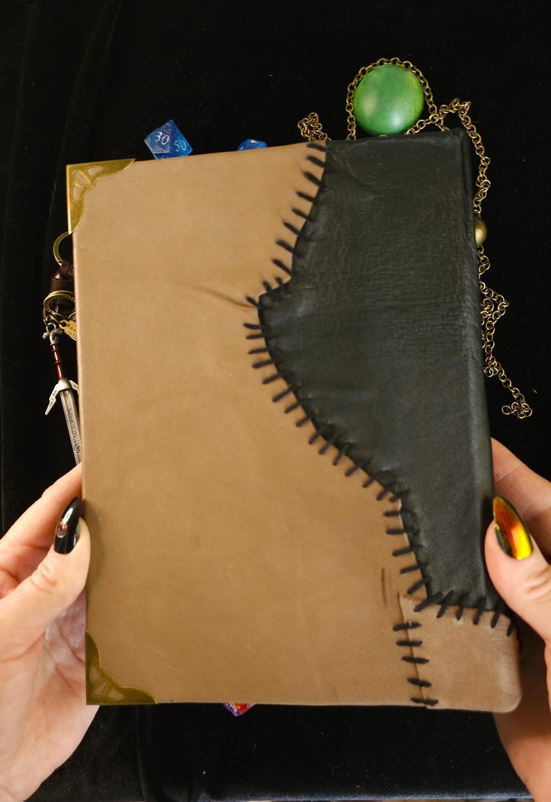 May include: A handmade leather journal with a two-tone design. The journal features a tan cover with a black leather accent stitched together with black thread. Decorative metal corner pieces and a sword charm are visible.