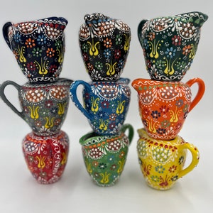May include: A set of eight small ceramic creamer pitchers with colorful hand-painted floral designs. The pitchers are stacked in a pyramid shape, with the largest pitcher at the bottom and the smallest pitcher at the top.