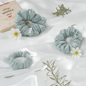 May include: Three light blue fabric scrunchies with white labels. The scrunchies are on a white surface with green sprigs.
