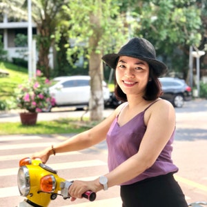 May include: A woman wearing a black hat and a purple tank top is riding a yellow moped. The moped has a white seat and a black rack on the back.