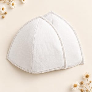 Puede incluir: Dos copas de sujetador triangulares de color blanco, hechas de un tejido texturizado con costuras visibles en los bordes. Las copas están colocadas una al lado de la otra y presentan una costura central que crea una forma redondeada.