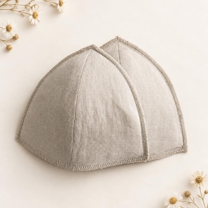 May include: A pair of triangular bra cups made from a light beige, textured fabric. Each cup features a structured, curved shape with visible stitched edges along the perimeter, providing a clean finish to the fabric construction.