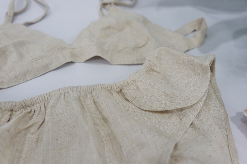 May include: A beige linen bralette and matching shorts with a ruffled trim. The bralette has adjustable straps.