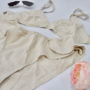 May include: A beige linen bikini top and bottom with ruffled trim. The top has adjustable straps and a triangle shape. The bottom has a high-waisted fit and a ruffled edge.