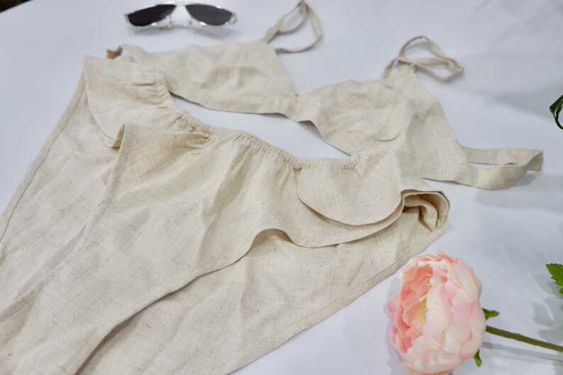May include: A beige linen bikini top and bottom with ruffled trim. The top has adjustable straps and a triangle shape. The bottom has a high-waisted fit and a ruffled edge.