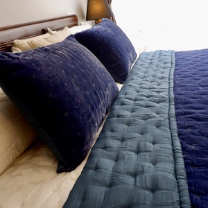 May include: A bed with a blue velvet quilted comforter and two blue velvet pillows. The bed has a wooden headboard and a white sheet.