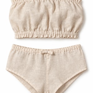 Puede incluir: Un conjunto de dos piezas en color beige que consta de un top tipo tubo sin tirantes con escote y dobladillo elásticos fruncidos, y pantalones cortos a juego con cintura elástica y un pequeño lazo decorativo centrado en la parte delantera.