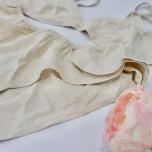 May include: A white linen lingerie set with a ruffled trim. The set includes a bra and a matching bottom. A pink flower is in the foreground.