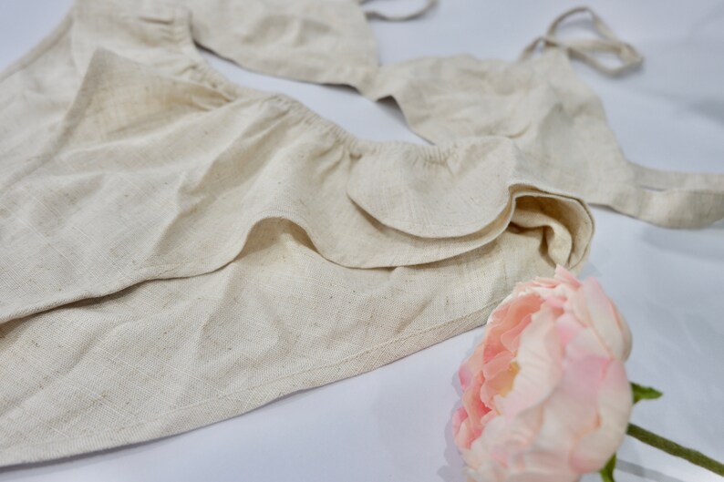 May include: A white linen lingerie set with a ruffled trim. The set includes a bra and a matching bottom. A pink flower is in the foreground.