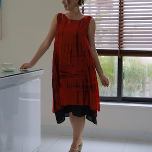 Puede incluir: Vestido rojo sin mangas con estampados tie-dye negros y una enagua negra. El vestido llega a la rodilla y se lleva con sandalias de cuña beige. El fondo incluye una ventana con persianas y una encimera blanca.