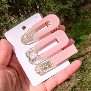 May include: Three pink hair clips with gold glitter accents. The clips are on a white card.