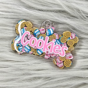 Puede incluir: Una etiqueta transparente en forma de hueso para mascotas llena de galletas pequeñas y redondas y dijes de cartón de leche. La palabra "Cookies" está escrita en rosa, con letras de burbujas. La etiqueta tiene un anillo plateado para sujetar.
