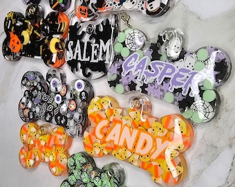 May include: A collection of eight Halloween-themed dog tags. The tags are shaped like bones and are decorated with various Halloween-themed designs, including ghosts, pumpkins, candy corn, and bats. The tags also have the names "Salem", "Casper", and "Candy" written on them.