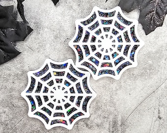 May include: Two white resin coasters with a black spiderweb design and iridescent glitter embedded in the resin.