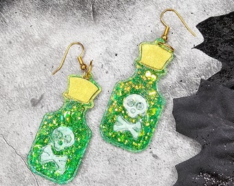 Peut inclure: Une paire de boucles d'oreilles en forme de fiole de potion verte à paillettes avec des sommets dorés et des motifs de crâne et d'os croisés.