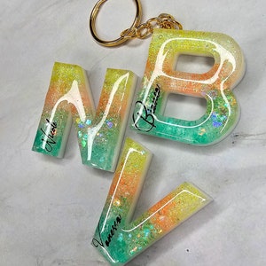 Könnte beinhalten: Ein Satz aus drei bunten Buchstabenanhängern aus Harz mit Glitzer, der in das Harz eingebettet ist. Die Buchstaben sind "N", "B" und "V" und jeder Buchstabe hat einen Namen darauf geschrieben. Die Buchstaben sind an einem goldenen Schlüsselanhänger befestigt.