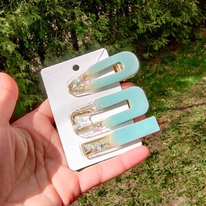 May include: Three teal and gold hair clips with glitter accents. The clips are on a white card.
