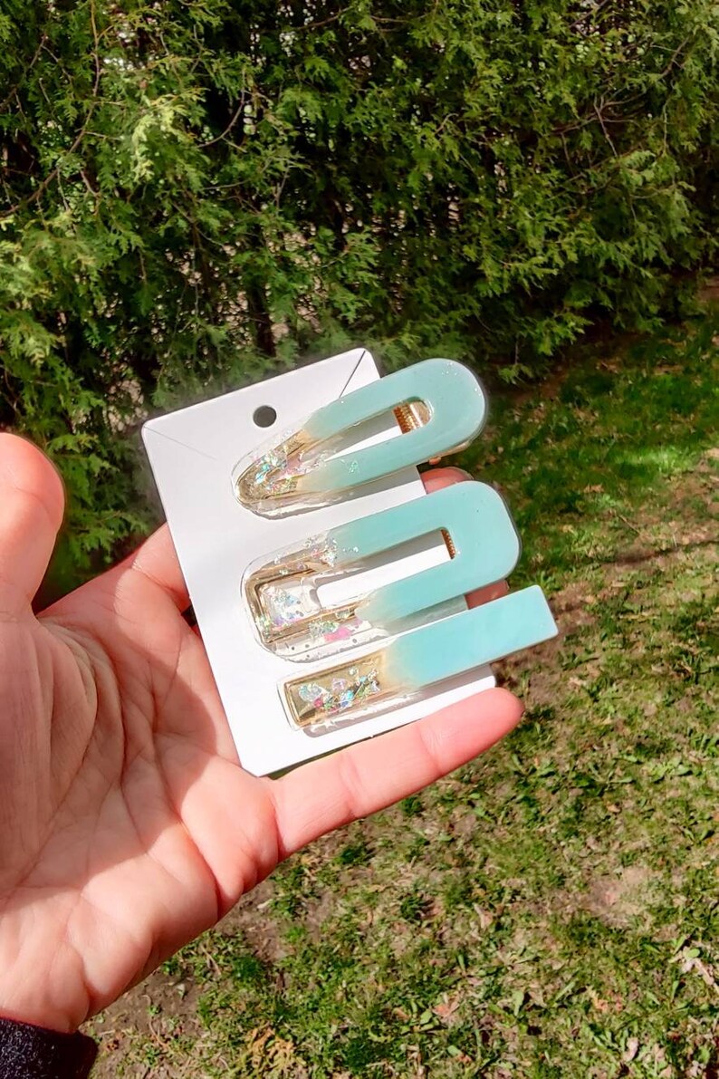 May include: Three teal and gold hair clips with glitter accents. The clips are on a white card.