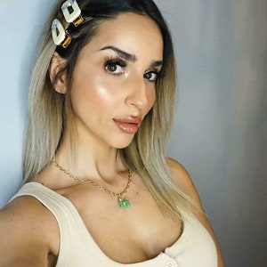 May include: A woman with long blonde hair wearing a white vest top and a gold chain necklace with a green butterfly pendant. Her hair is pulled back with two white and gold hair clips.