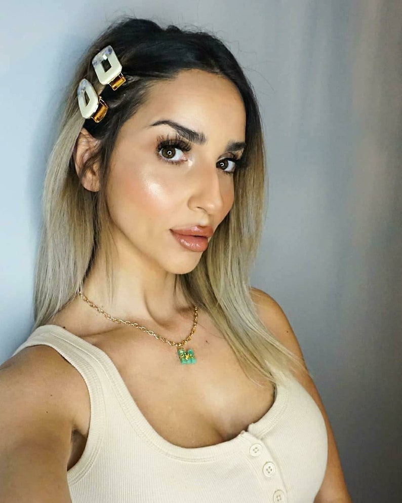 May include: A woman with long blonde hair wearing a white vest top and a gold chain necklace with a green butterfly pendant. Her hair is pulled back with two white and gold hair clips.