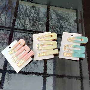 May include: Three sets of hair clips with a gradient colour design. Each set has three clips in different sizes. The clips are decorated with gold glitter. The clips are on a white background.