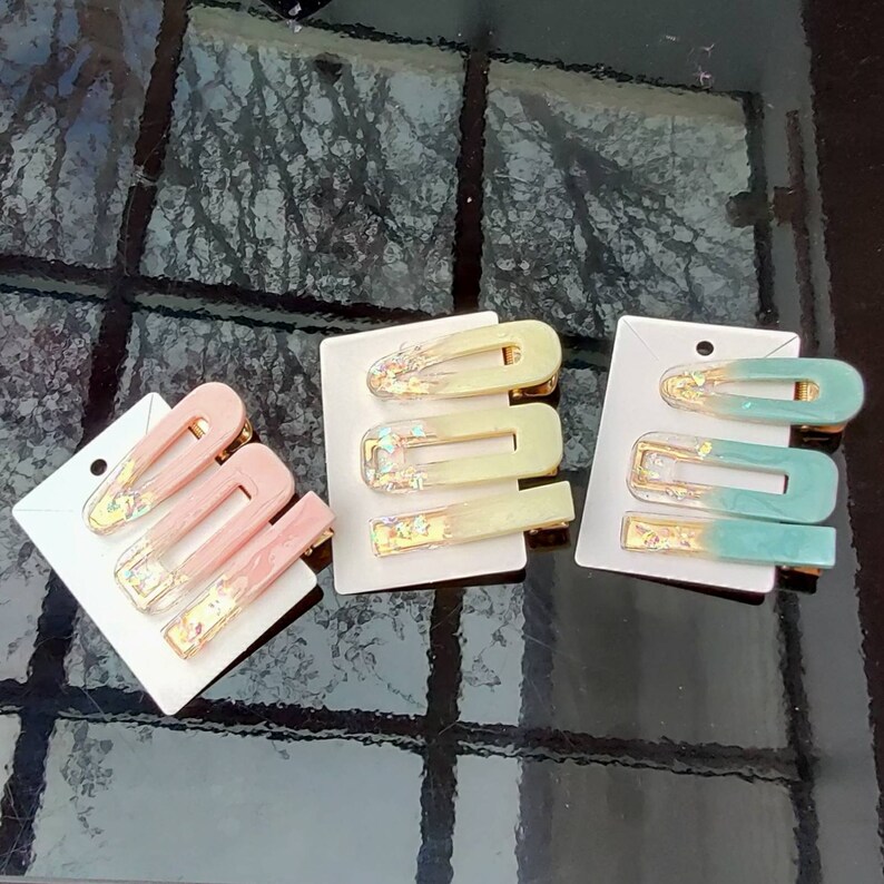 May include: Three sets of hair clips with a gradient colour design. Each set has three clips in different sizes. The clips are decorated with gold glitter. The clips are on a white background.