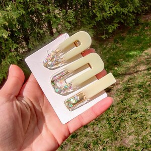 May include: Set of three hair clips with a pale yellow translucent base and iridescent glitter embedded inside. The clips are on a white card.