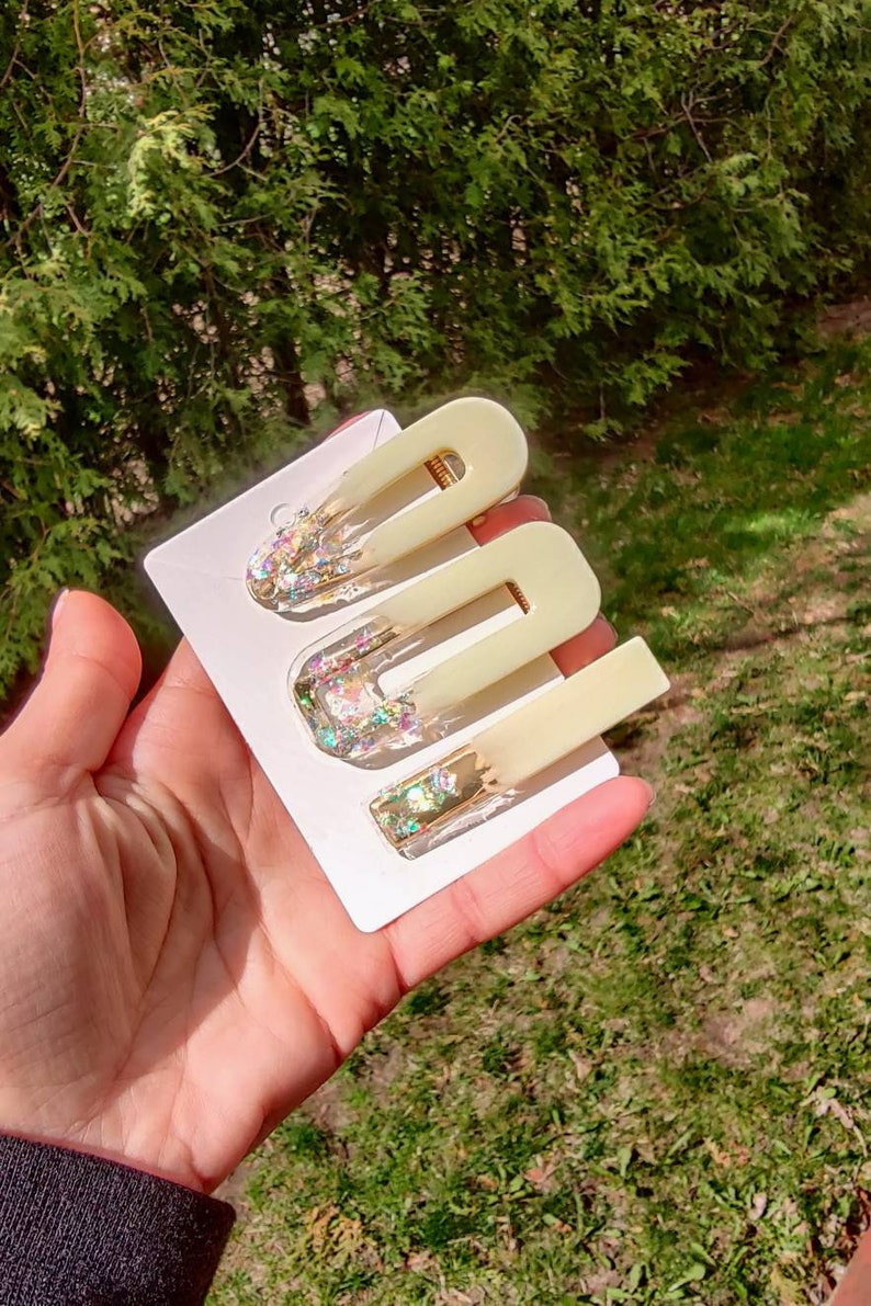 May include: Set of three hair clips with a pale yellow translucent base and iridescent glitter embedded inside. The clips are on a white card.