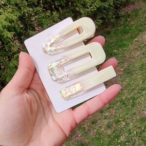 May include: Set of three white hair clips with gold accents and iridescent glitter. The clips are in different sizes and shapes.