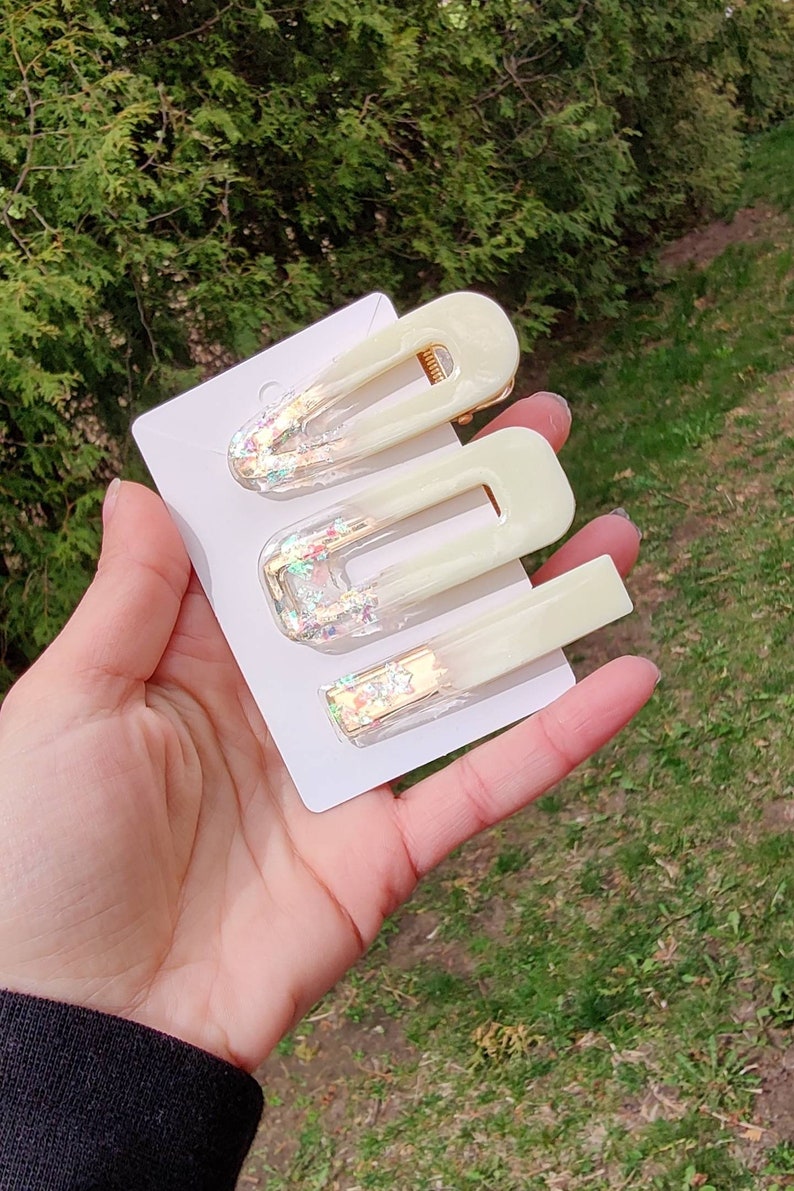 May include: Set of three white hair clips with gold accents and iridescent glitter. The clips are in different sizes and shapes.