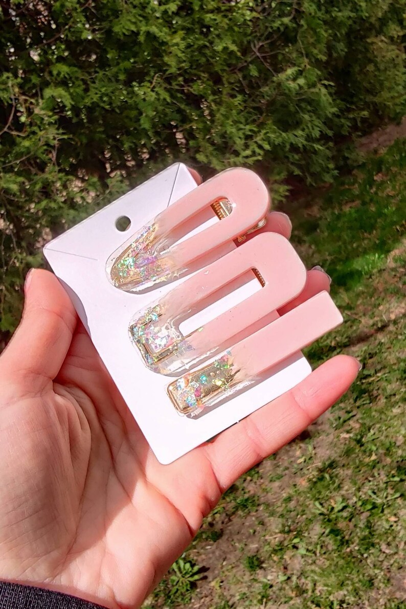 May include: Three pink hair clips with gold glitter accents. The clips are on a white card.