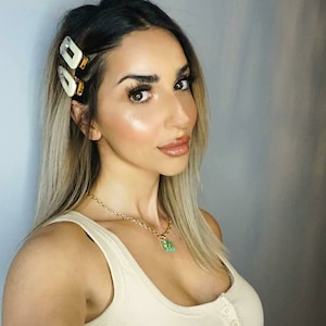 May include: A woman with long blonde hair wearing a white tank top and a gold chain necklace with a green pendant. She has a pair of white hair clips in her hair.