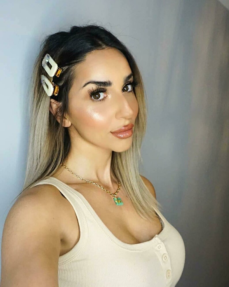 May include: A woman with long blonde hair wearing a white tank top and a gold chain necklace with a green pendant. She has a pair of white hair clips in her hair.