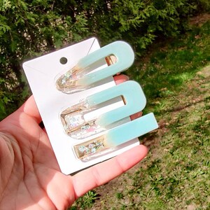 May include: Set of three blue and gold hair clips with glitter accents. The clips are in the shape of a U and are arranged on a white card.