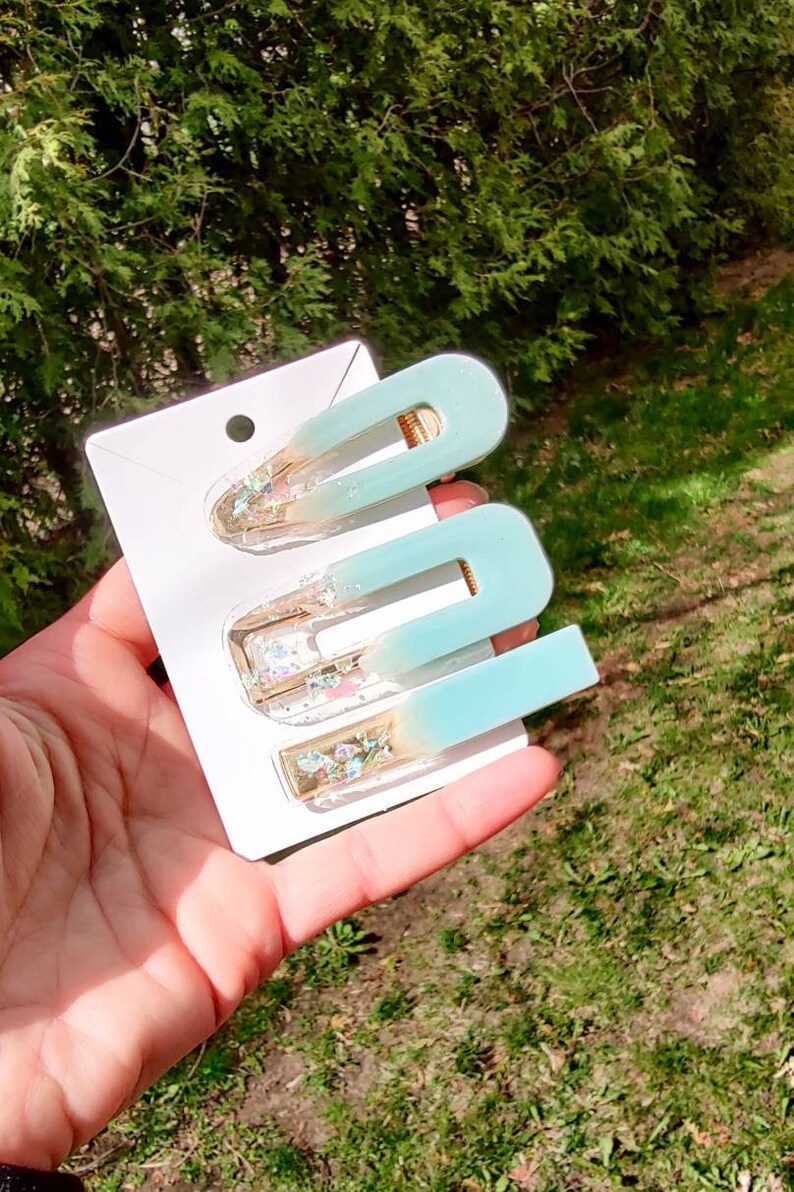 May include: Set of three blue and gold hair clips with glitter accents. The clips are in the shape of a U and are arranged on a white card.