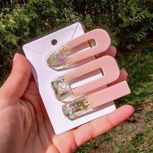 May include: Three pink hair clips with gold glitter accents. The clips are on a white card.